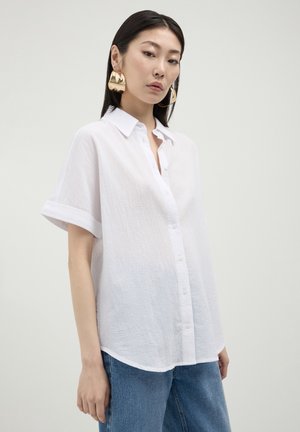 Donna che indossa una camicia button-up bianca oversize e testurizzata a maniche corte, jeans blu e grandi orecchini d'oro audaci su uno sfondo semplice.