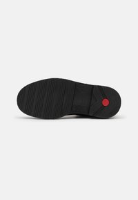Zwarte rubberen sneaker zool met een getextureerd patroon, voorzien van een rode cirkelvormige accent en horizontale looplijnen voor grip.