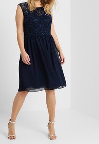 Robe marine avec un corsage en dentelle et une jupe en chiffon fluide, présentant une taille ajustée et un design sans manches. Sandales à talons hauts argentées.