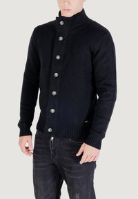 Uomo che indossa un cardigan a maglia nero con bottoni e colletto alto, abbinato a jeans neri, su uno sfondo chiaro e uniforme.