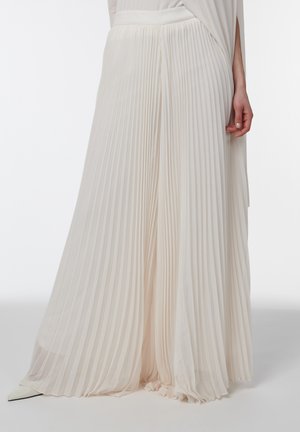 Pantalon blanc plissé, ample et à jambes larges, porté par une personne debout, accompagné de chaussures blanches pointues, sur un fond clair uni.