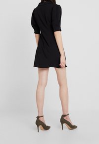 Black short-sleeve dress with a collar and buttoned cuffs, featuring a fitted silhouette. Paired with olive green high-heeled shoes.