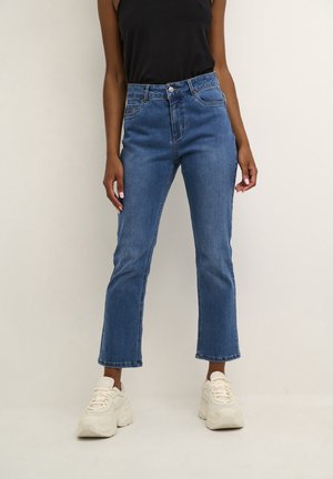 Persoon die blauwe cropped flare jeans, een zwart mouwloos topje en witte chunky sneakers draagt, staand tegen een effen achtergrond.
