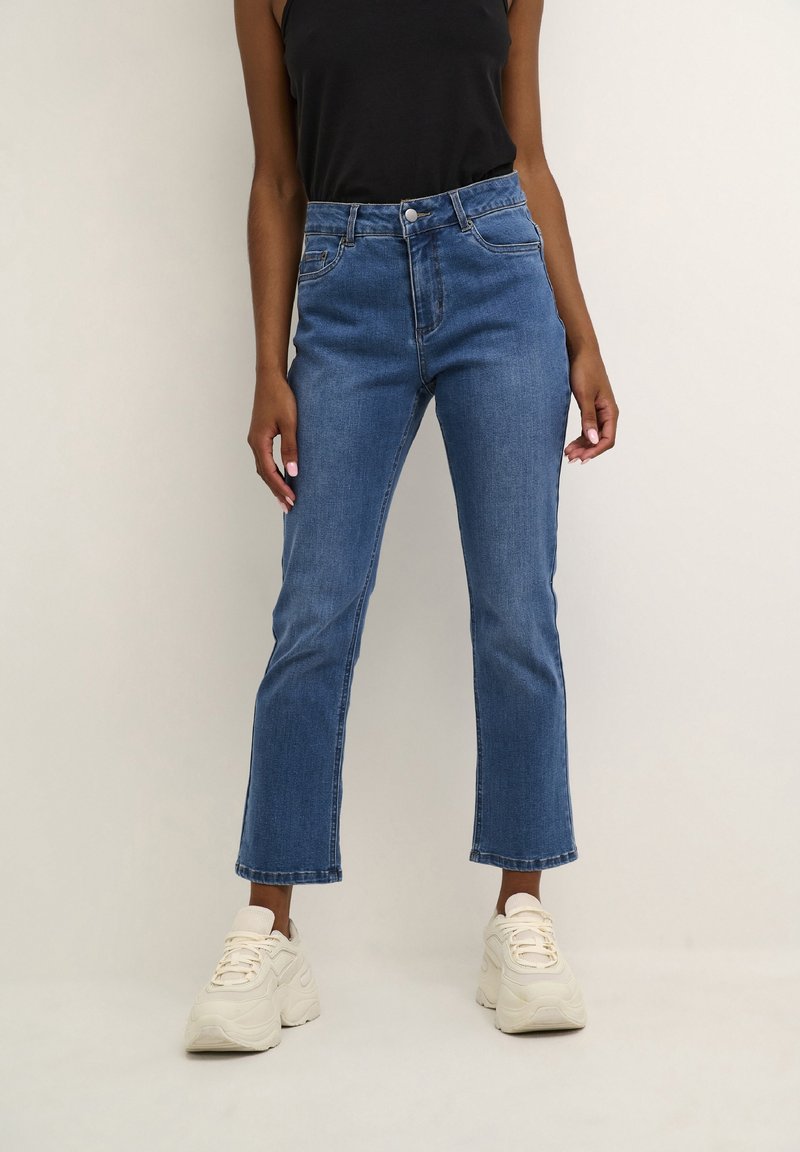 Personne portant un jean flare court bleu, un haut noir sans manches et des baskets blanches épaisses, debout devant un fond uni.