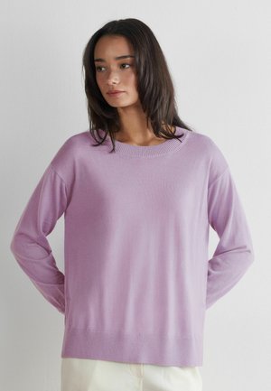 Jeune femme aux cheveux bruns mi-longs portant un pull ample lavande et un pantalon clair, debout les mains derrière le dos.