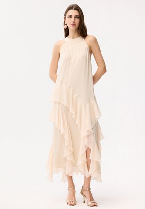 Kobieta stoi ubrana w bezrękawową, jasnobeżową, warstwową sukienkę midi z falbanami oraz złote szpilki na kostkę, na jednolitym białym tle.