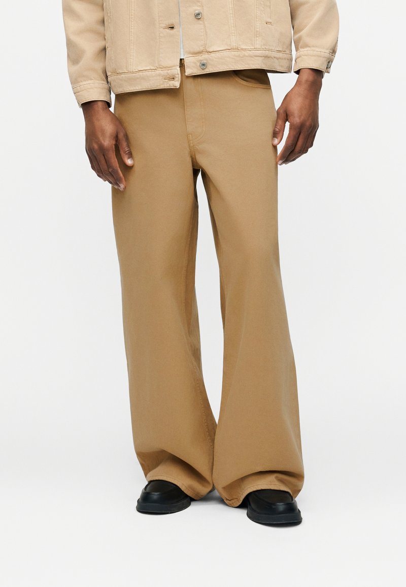 Άνδρας που φοράει μπεζ παντελόνι φαρδιάς γραμμής, μπεζ μπουφάν και μαύρα παπούτσια, πάνω σε απλό λευκό φόντο.