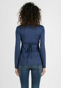 Marinefarbener Langarmoberteil mit einem gebundenen Design im Rücken, das eine glatte Textur aufweist. Kombiniert mit blauen Denim-Jeans.