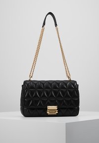 Černá prošívaná kožená kabelka se zlatým řetízkem a obdélníkovou zlatou sponou na bílé výstavní ploše.