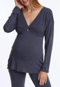 Zwangere top in donkergrijze stof met lange mouwen, V-hals en knoopdetail bij de borst. Gladde textuur, losse pasvorm, met logo-accent op de mouw.