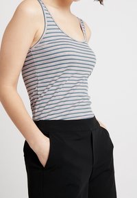 Top de tirantes a rayas en gris claro, azul y rosa. Presenta un diseño ajustado con tirantes finos y una textura suave. Combinado con pantalones negros.