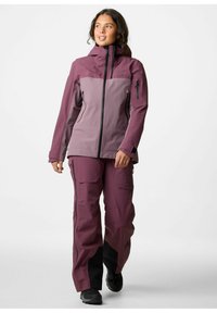 Veste et pantalon de ski violets, avec un panneau supérieur contrastant de couleur plus claire, des poches zippées et un tissu texturé, avec des accents noirs sur le pantalon.