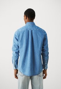 Camisa de mezclilla azul con cuello, puños con botones y dobladillo curvado. La tela tiene una apariencia texturizada, diseñada para el uso casual.