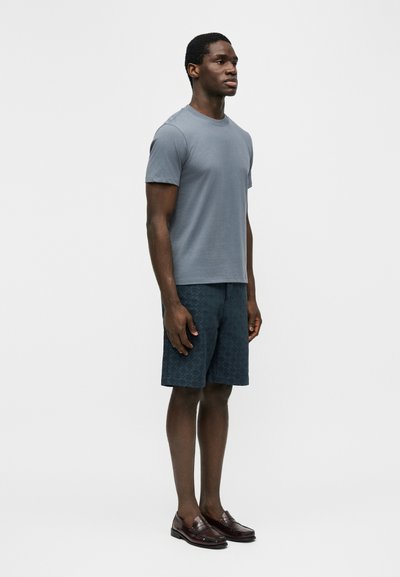 Άνδρας που στέκεται φορώντας γκρι μπλουζάκι, σκούρα σορτς με μοτίβο και καφέ δερμάτινα μοκασίνια μπροστά σε απλό λευκό φόντο.