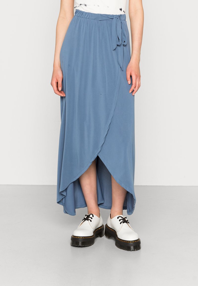 Jupe midi bleue portefeuille avec détail de nœud sur le côté portée avec des chaussures à lacets blanches à plateforme sur une personne debout.