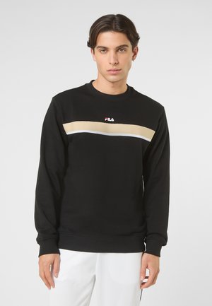 Junger Mann mit kurzem dunklem Haar, der einen schwarzen FILA-Sweatshirt mit beigen und weißen horizontalen Streifen trägt, kombiniert mit weißen Hosen.