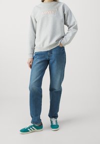 Persona con un sudadera gris de Levi’s, jeans azules y zapatillas teal de Adidas con rayas blancas, de pie con una mano en el bolsillo.