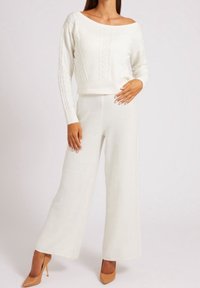 Weißer Strickpullover mit schulterfreiem Design und Zopfmuster, kombiniert mit weit geschnittenen weißen Hosen aus weichem Material und minimaler Gestaltung.