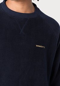 Felpa di pile navy con colletto a costine, caratterizzata da un piccolo logo dorato "woodbird." sul petto. Texture morbida e design casual.