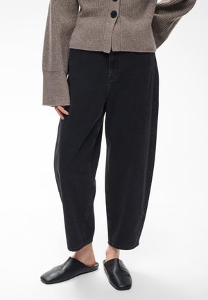 Personne portant un pantalon court noir ample, des chaussures noires sans lacets, et un pull en tricot marron boutonné à manches longues.