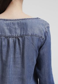 Henkilö, joka näkyy selkäpuolelta, pukeutuneena siniseen farkkupaitaan, jossa on kokoontunutta ommelta yoke-alueen alla, yksinkertaisella vaaleanharmaalla taustalla.