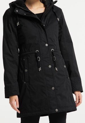 Parka longue noire à capuche avec boutons sur le devant, cordons de serrage réglables à la taille et poches, portée sur des vêtements noirs.