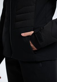 Svart vadderad jacka med dragkedja, textil med struktur och en sidoficka med kardborre. Slimmad passform med en slät yta och minimalistisk design.