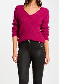 Frau trägt einen leuchtend pinken V-Ausschnitt-Pullover und hochtaillierte schwarze Hosen mit dekorativen goldenen Knöpfen an den Taschen und dem Bund.