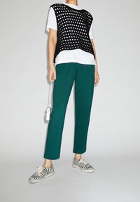 Un débardeur en maille noire sur un t-shirt blanc, associé à un pantalon teal et des sneakers à motif zèbre. Tient un appareil avec une corde.