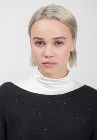 Zwarte trui met een ruime halslijn, versierd met kleine zilveren accenten, gelaagd over een witte coltrui. Lichtblond haar. Neutrale achtergrond.