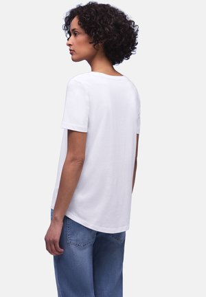 Vrouw met krullend haar in zijaanzicht, gekleed in een effen wit T-shirt met korte mouwen en blauwe spijkerbroek, tegen een witte achtergrond.