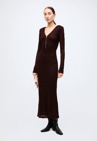 Long-sleeve burgundy dress with a lace pattern, V-neckline, and button detail. Fitted silhouette, floor-length, paired with black boots.