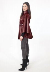 Capa de cuero sintético burdeos con cuello alto y cintura con cinturón, combinada con jeans ajustados negros y botines de tacón stiletto. Textura suave.