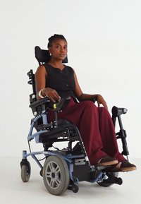 Ηλεκτρικό αναπηρικό καροτσάκι με μπλε σκελετό, μαύρη θέση και μεγάλους γκρίζους τροχούς. Ο χρήστης φοράει μαύρη αμάνικη μπλούζα και μπορντώ παντελόνι. Απλή σχεδίαση.