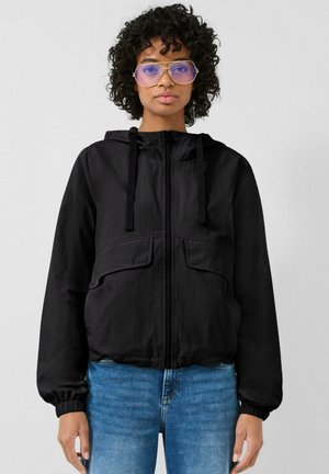 Kobieta o kręconych włosach, nosząca duże fioletowe okulary, czarną bluzę z kapturem na suwak i niebieskie dżinsy, stojąca na jednolitym tle.