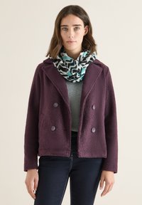 Purple fleece jacket with a textured surface, double-breasted design, and large buttons. Worn over a grey sweater, accompanied by a patterned scarf.