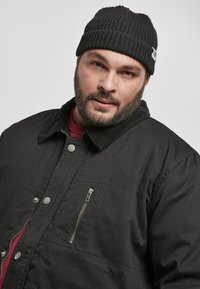 Giacca nera trapuntata con colletto e tasca sul petto con zip. Indossata con un berretto nero a coste. Camicia rossa visibile sotto.