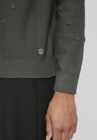 Ciemnoszary sweter z dzianiny z teksturowanymi akcentami w postaci guzików, ściągaczami na mankietach i dole, z małą metalową metką z logo blisko dołu.