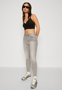 Crna kratka majica s prednjim twistom, uparena s visokim svjetlosivim skinny trapericama i srebrnim tenisicama. Elegantne, moderne naočale naglašavaju izgled.