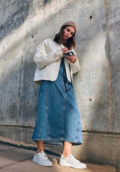Bijela quilted jakna, plava denim midi haljina i bijele tenisice. Braon pleteni šešir i vintage kamera u ruci. Pozadina: betonski zid.