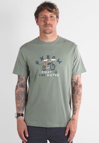 T-shirt en coton vert olive avec un graphisme représentant un surfeur à vélo et le texte "OXBOW FIRST IN THE WATER." Manches courtes, col rond.