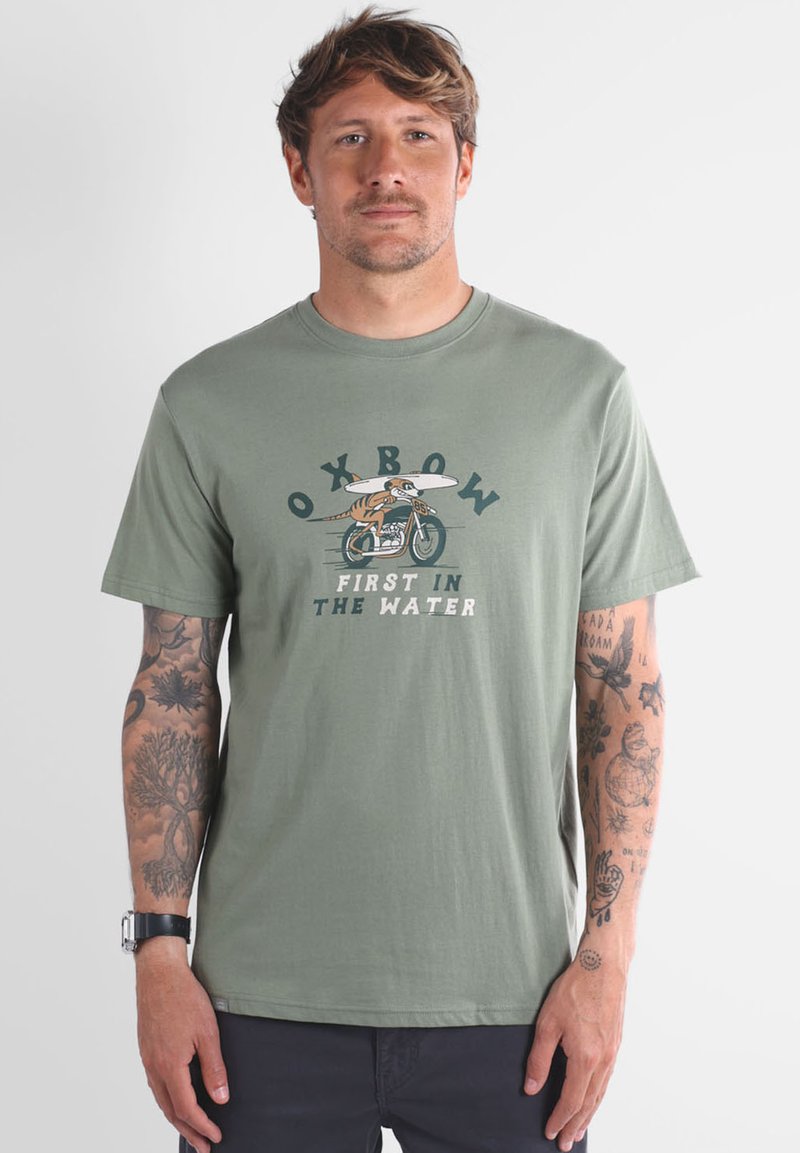 T-shirt en coton vert olive avec un graphisme représentant un surfeur à vélo et le texte "OXBOW FIRST IN THE WATER." Manches courtes, col rond.