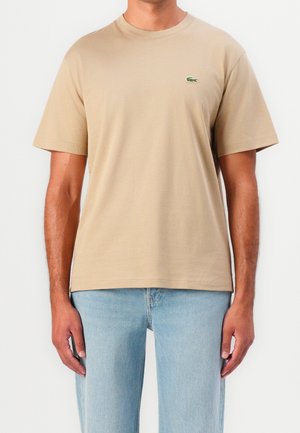 Homme portant un t-shirt beige à manches courtes avec un logo de crocodile vert sur la poitrine gauche et un jean bleu clair.