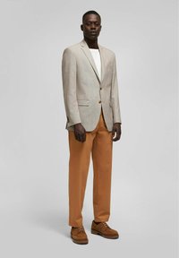 Hellgrauer Blazer mit zwei Knöpfen und Reverskragen, kombiniert mit rostfarbenen Hosen und braunen Schnürschuhen. Darunter ein schlichtes weißes T-Shirt.