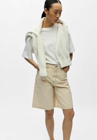 Femme aux cheveux noirs portant un t-shirt blanc, un pull blanc drapé sur les épaules et un short beige longueur genou, les mains dans les poches.