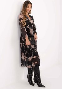 Schwarzes plissiertes Kleid mit hellrosa Blumenmustern, langen Ärmeln und V-Ausschnitt. Kombiniert mit kniehohen schwarzen spitzen Stiefeln und einer Clutch.