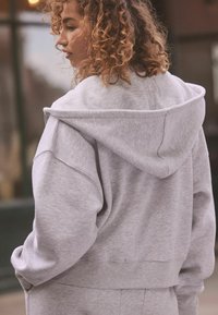 Felpa grigia con cappuccio, di vestibilità rilassata, con polsini e vita a costine. Presenta una zip e una texture morbida, con linee e cuciture curve.
