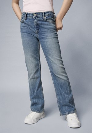 Niebieskie dzwony z denimu o wyblakłej fakturze, z pięcioma kieszeniami i szlufkami na pasek. Noszone z białymi trampkami i jasnoróżową bluzką.