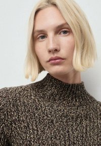 Donna bionda con capelli corti che indossa un maglione a collo alto a maglia mélange marrone e beige, espressione neutra, sfondo chiaro.