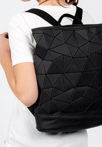 Svart ryggsäck med geometriskt mönster gjord av texturerat material, med justerbara remmar och ett handtag upptill. Har en slät svart bas.